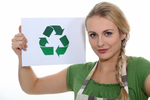 A young woman with blonde hair styled in a side braid, wearing a green short-sleeved top and a grey striped apron, is holding up a white sign with a large green recycling symbol on it. She is standing against a plain white background and has a neutral facial expression. The recycling symbol consists of three arrows forming a continuous loop, symbolising waste recycling and environmental sustainability, which aligns with waste management services. The image emphasizes eco-friendly themes relevant to rubbish and waste removal in the Chigwell area, and the woman’s attire suggests involvement in environmental or waste disposal work. The focus on the recycling sign and the woman’s confident stance underscores the importance of waste management and recycling initiatives, supporting the professional image of [COMPANY_NAME] offering rubbish removal services in the vicinity of Chigwell, Essex, without depicting any specific rubbish objects or waste materials.