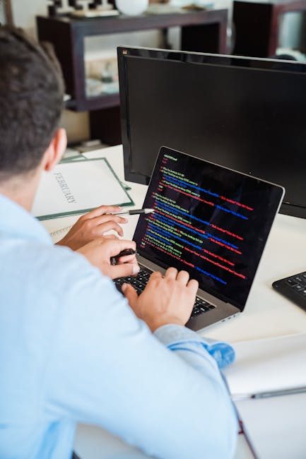 A person working at a desk with two computer monitors and a laptop, one monitor displaying lines of colorful computer code on a dark background. The individual, wearing a light blue shirt, is seated w