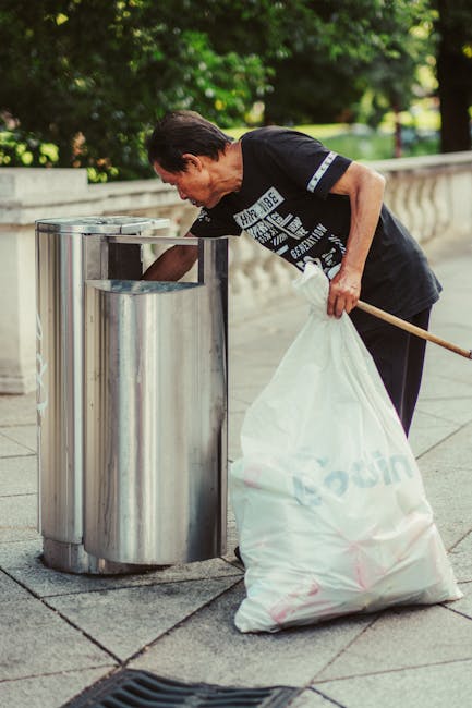A person wearing a short-sleeved shirt is seen disposing of paper waste by placing a crumpled brown paper bag into a grey waste bin with a label indicating it is for rubbish. The bin is situated on a 